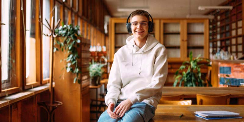 Young male student sitting at library desk wearing a white sweater and headphones smiling