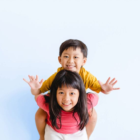 Young student in a yellow shirt climbing on the back of a young student in a red shirt  with their arms raised in front of a blue wall