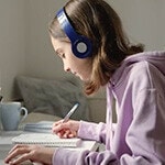 A middle school student taking notes and listening to an online class through headphones