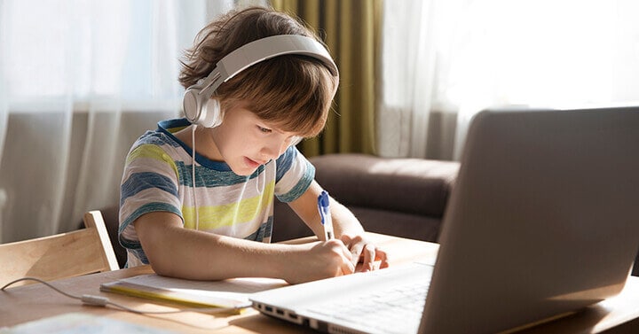 Young male student writes on paper in front of a laptop while wearing headphones
