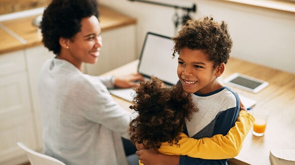 A mother is working on a laptop while her son and daughter hug