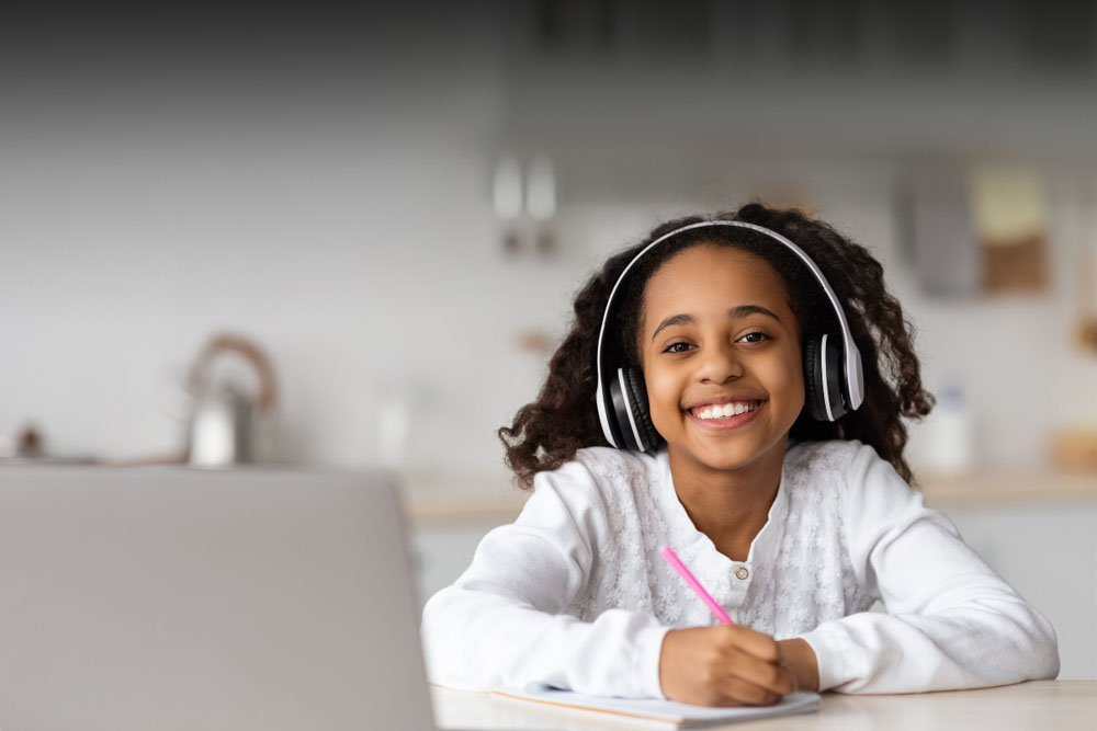 Girl in a white shirt looking at the camera smiling while writing in her school journal taking an online class at Inspire Connections Academy. 