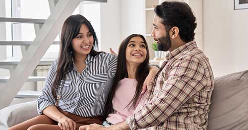 An image of a girl sitting on the sofa with her mother and father and looking up smiling at her father.