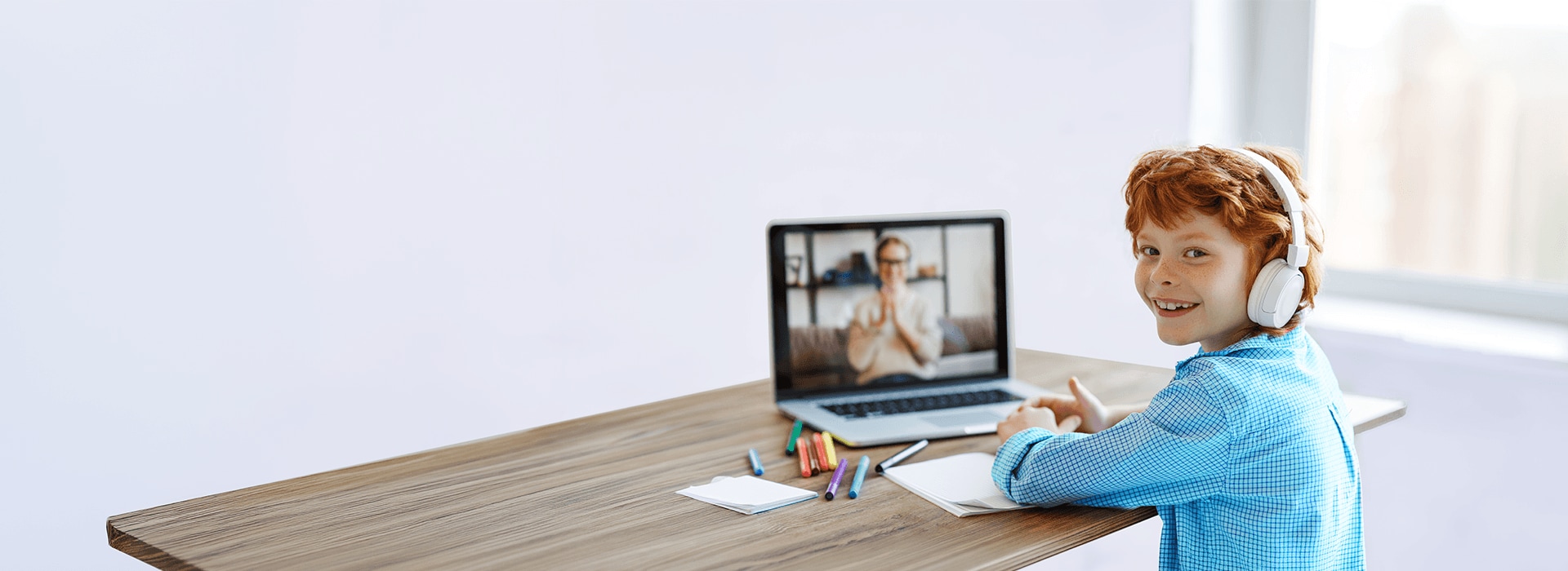 Smiling student wearing white headphones sits at a desk with notebooks and markers, attending an online video lesson on a laptop.