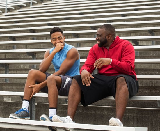 A high school athlete in a blue shirt talks with his coach in a red sweater sitting on bleachers during a track and field practice. 