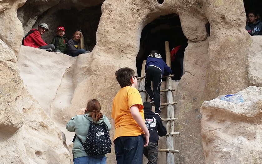 People explore ancient cave dwellings, climbing a wooden ladder and standing in carved openings, in a rocky outdoor setting.
