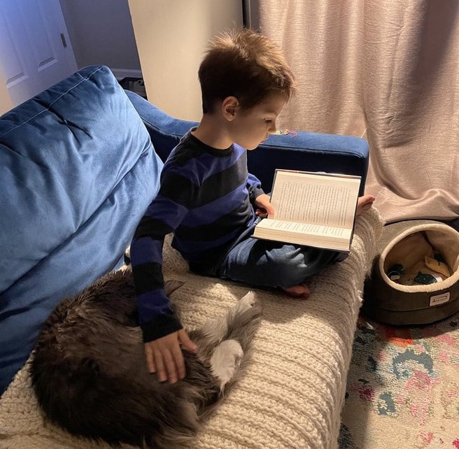 Leos sitting on a couch reading a book while petting his cat. 