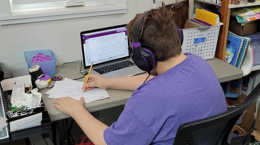 Lucus sitting at his desk and writing on a piece of paper with his school laptop open. 