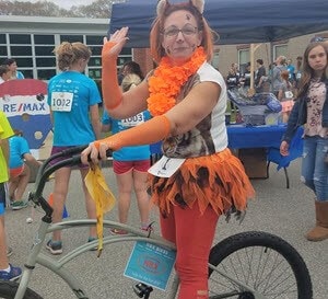 Tracey waving on a bike at an event while dressed in an orange costume