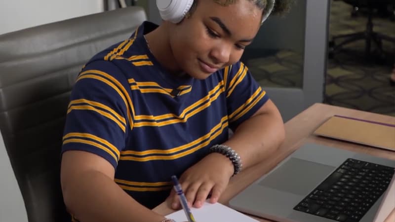 An online school student sitting in front of a laptop and wearing headphones and writing something down on paper