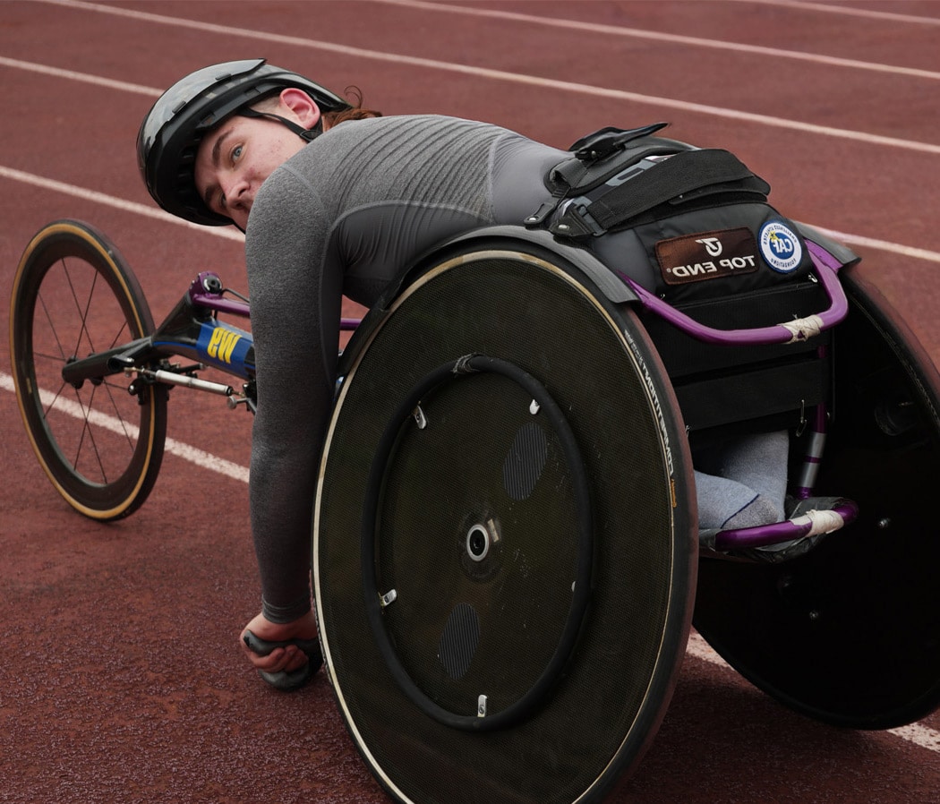 A student para-athlete in a racing wheelchair leans forward on a track, wearing a helmet and preparing to push off for a race.