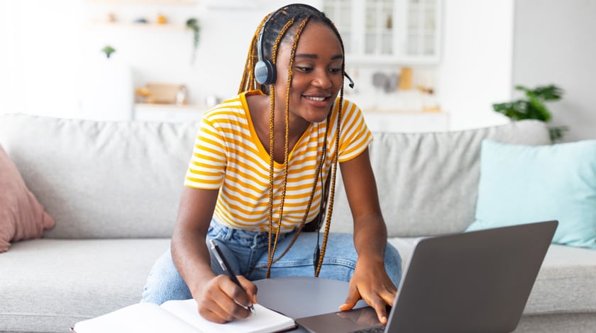Online school student sitting on couch, wearing headphones, writing in a notebook and using laptop