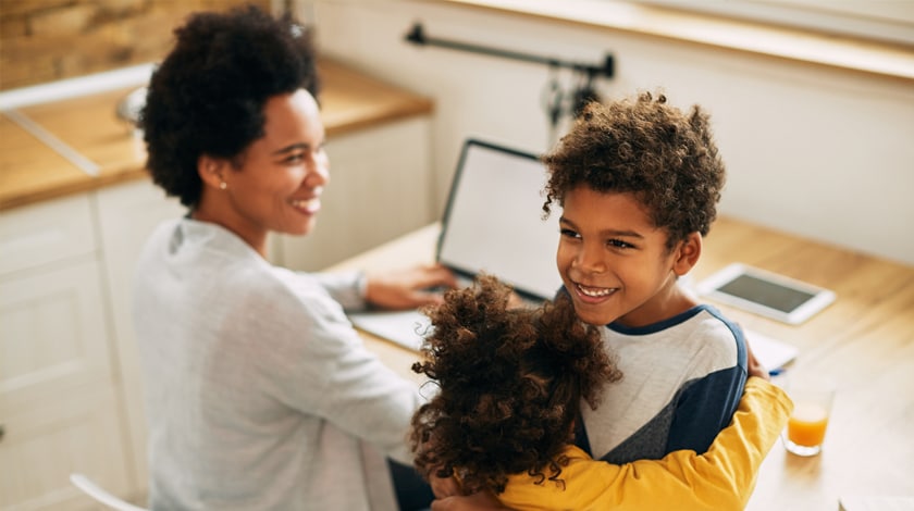 A mother is sitting at a computer while her son and daughter hug eachother