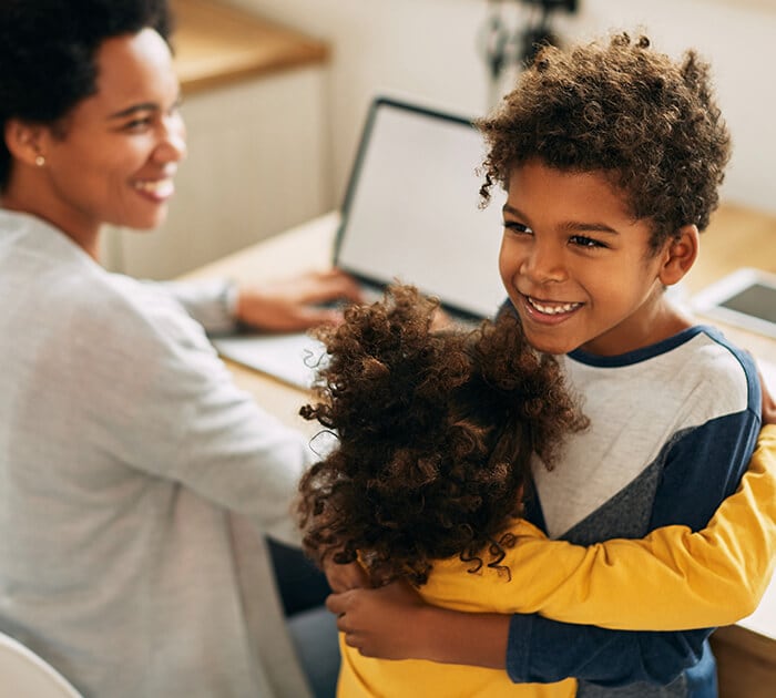 A brother and sister are hugging while their mom is on a laptop