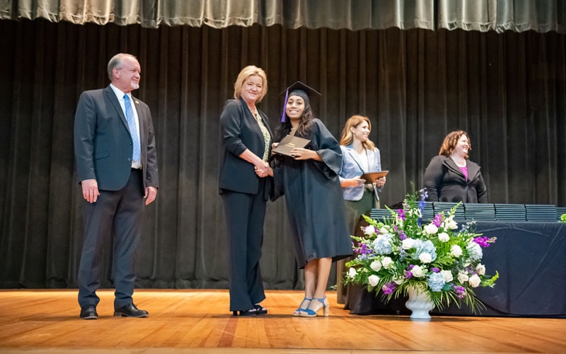 A graduating student in a black cap and gown receives her diploma on stage from Ms. Jodie Dean. A man in a suit stands to the side, and there is a table with more diplomas and a floral arrangement on stage.