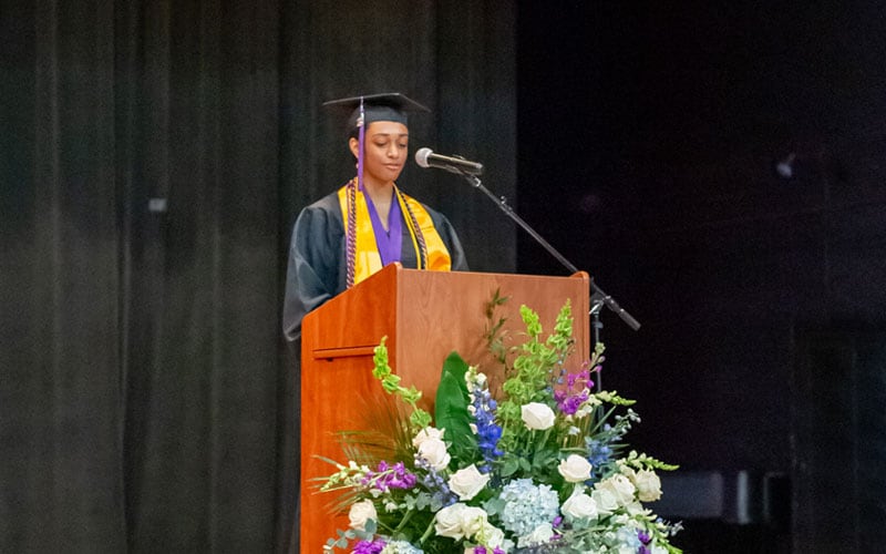 A graduate in a cap and gown stands at a podium decorated with flowers, delivering a speech at the Alabama Connections Academy graduation ceremony.