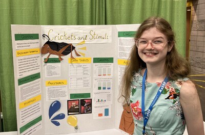 Image of Lilyana P presenting at the Arizona Science and Engineering Fair. She is wearing a blue dress and is presenting her project on Crickets and Stimuli.