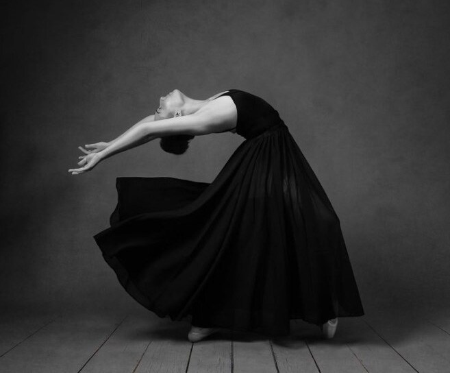 Black and white image of Ashlyn Hall in a black flowing dress with her arms thrown back arched behind her while on pointe. 