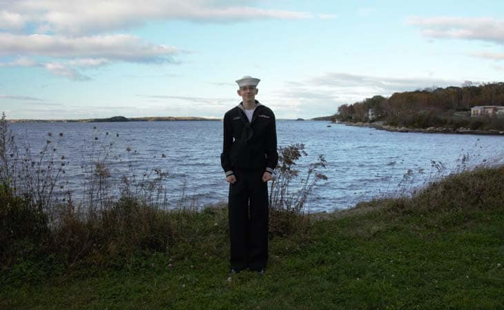 A person in a naval uniform stands by a lake with distant hills and sparse vegetation under a cloudy sky.
