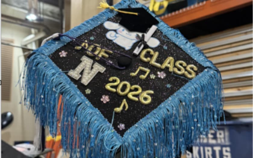 Decorated graduation cap with blue fringe; black glitter top shows a white long‑eared cartoon in a tiny mortarboard, gold letters “CLASS” and “2026,” a large silver “N,” small flowers, and musical notes.