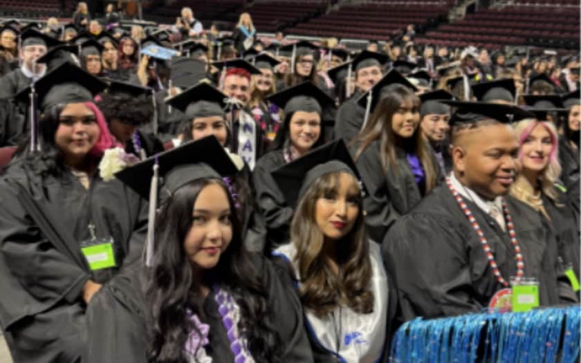 Rows of people in black graduation caps and gowns seated in an arena; faces are blurred. Some wear stoles, cords, or leis, and a shiny blue fringe decoration is at the lower right.