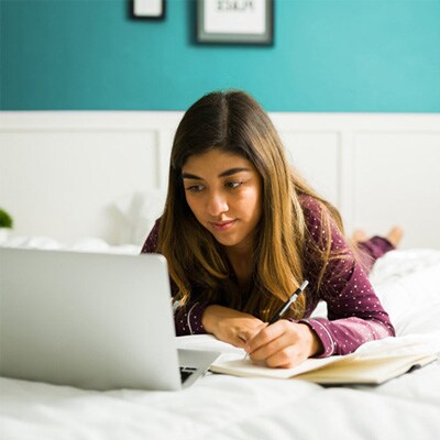 A teen student in an purple shirt sitting on her bed working on an online Geometry assigment.
