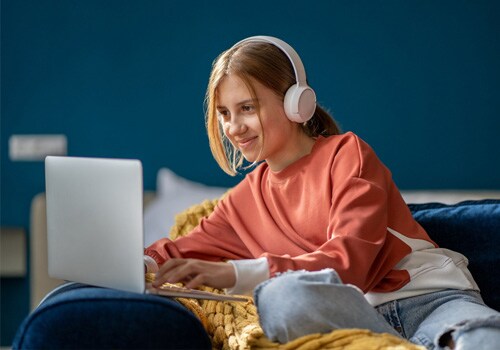 Teen female student in an orange t-shirt sitting against their bed working on an online assignment for Connections Academy.