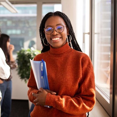 A young teen in an orange sweater smiling at the camera holding notebooks.  