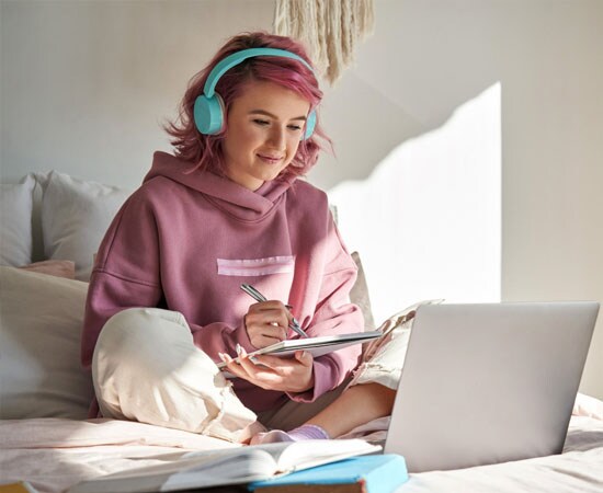 Connections Academy student in a pink sweat shirt sitting on her bed while smiling at her latpop.