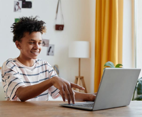 Connections Academy smiling student in a brown striped sweater shirt looking at his laptop while sitting at a desk working on an online assigment. 
