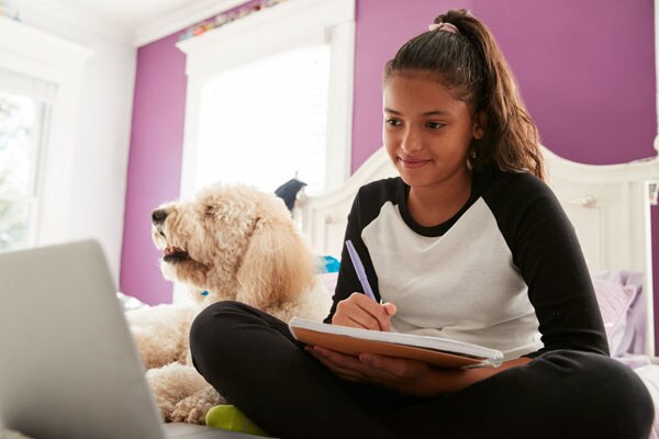 Image of a student in a black and white t-shirt sitting in their bed working on an online assignment. 
