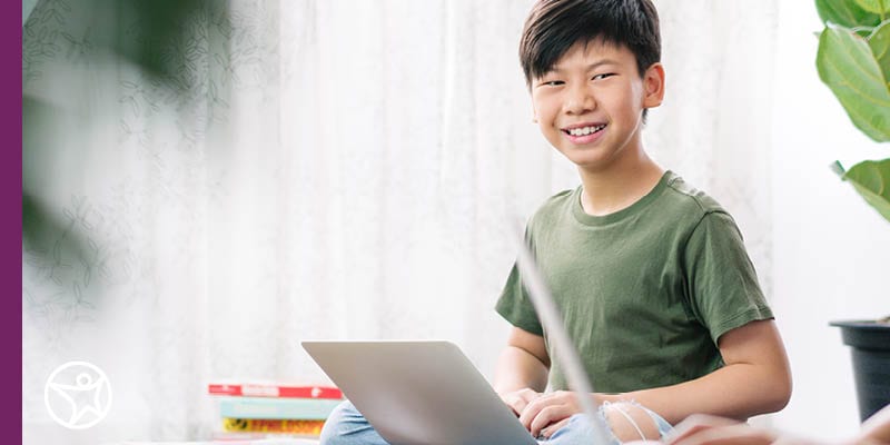 A middle school student working on his laptop sitting on the floor wearing a green t-shirt.