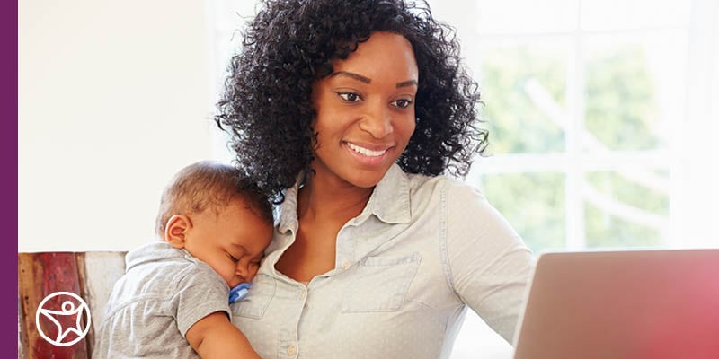 A woman holding a baby while working on a laptop at a table near a window.