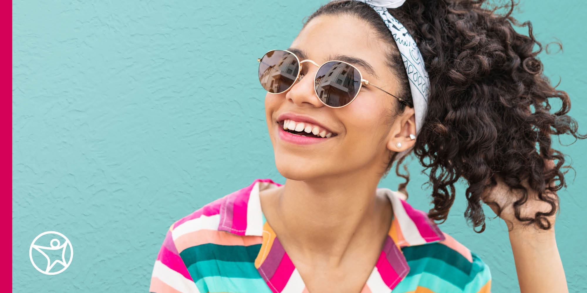 A student wearing sunglasses and a pink and blue striped shirt looking up and smiling against a blue wall.