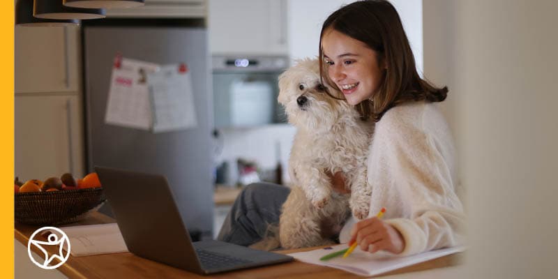 An online school student holding a small white dog and taking part in an online lesson on their laptop