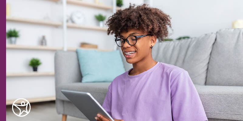 A Connections Academy online high school student sitting on the floor in front of a grey couch and working on their tablet.
