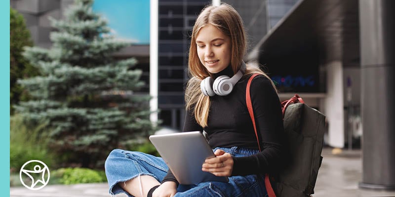 An online school student sitting outdoors with a tablet, wearing headphones around their neck and a backpack.