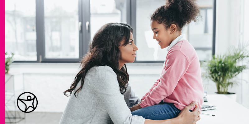A mother holds her daughter giving her a reassuring look and smile while she is sitting on a desk
