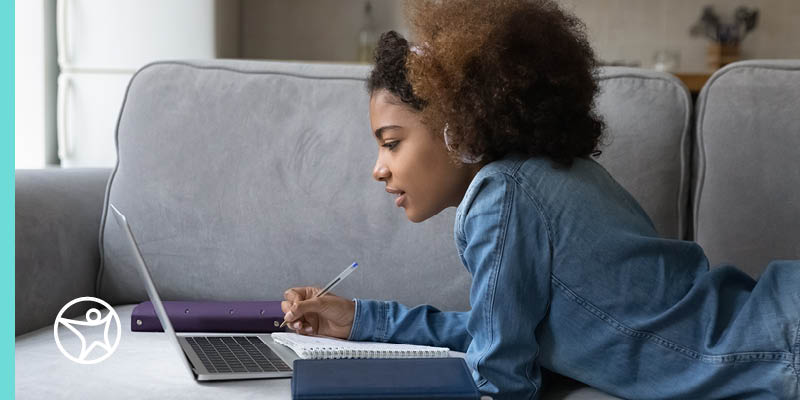 A teenage girl is laying on a couch with her laptop and taking notes