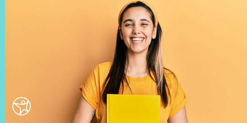 Student in a yellow shirt standing in front of a yellow wall holding a yellow folder smiling.