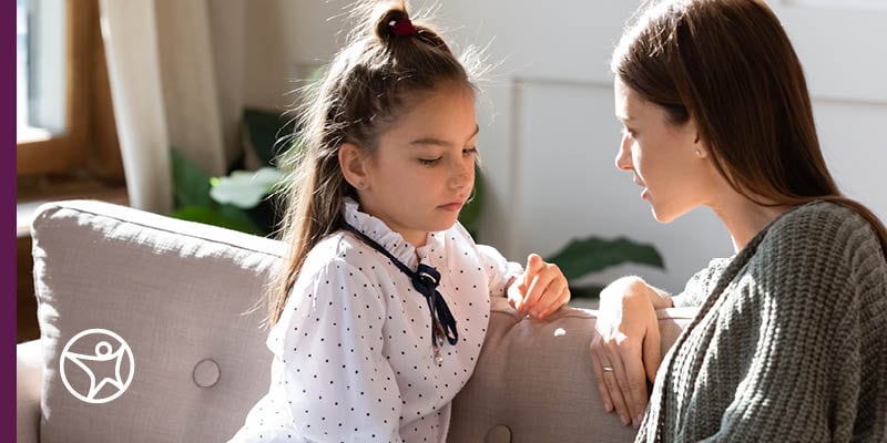 A mom is sitting on a couch and having a conversation with her young daughter