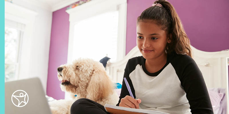 A student is sitting on her bed with dog while taking notes for an online class