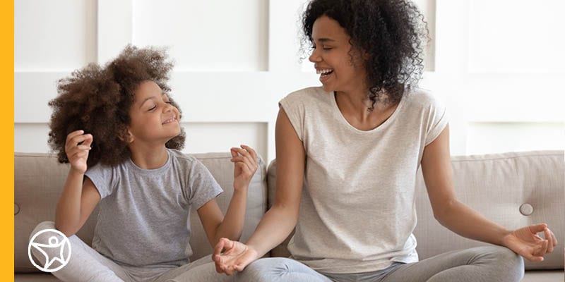 An adult and a child sitting on a couch in a relaxed pose, both wearing casual grey clothes and appearing to meditate to help manage stress.