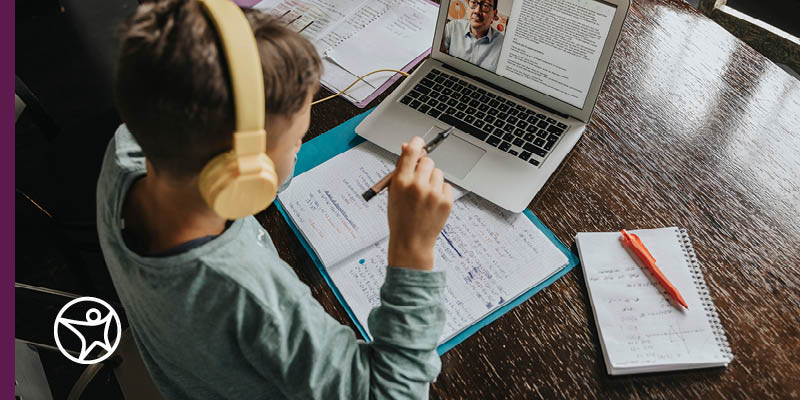 An elementary student wearing headphones and studying with a notepad and laptop on a desk