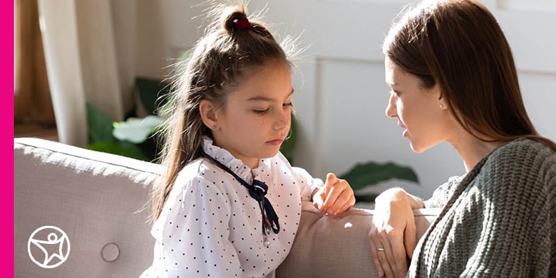 A mom wearing a grey sweater and sitting on a grey sofa talking to her young elementary age daughter wearing a polka dot with a black bow shirt also sitting on a grey sofa.