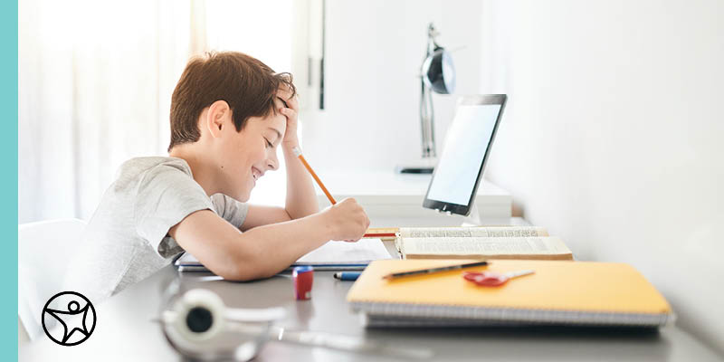 A young boy studing at a desk with his hand on his forehead while he smiles and writes somethig in a notebook.