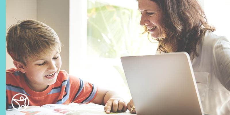 A mom wearing a white shirt smiling and looking at her young elementary age son wearing an orange and a blue striped shirt talking about an online assignment for Connections Academy.