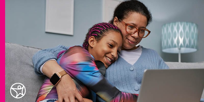 A mom in a blue sweater hugging her daughter while sitting on the sofa looking at a laptop making a plan to set goals at Connections Academy.
