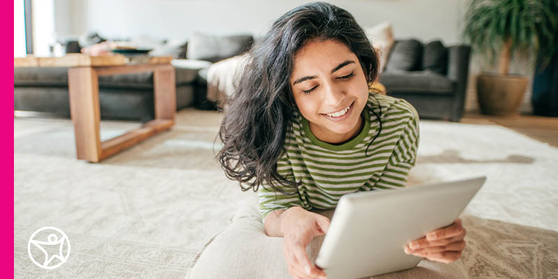 A young teen girl in a green striped shirt sitton on a white carpet rug looking at an IPAD for an online assignement at Connections Academy.