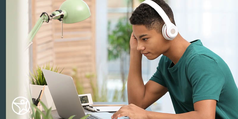 A high school student wearing a green shirt sitting at a desk working on a laptop with white headphones.